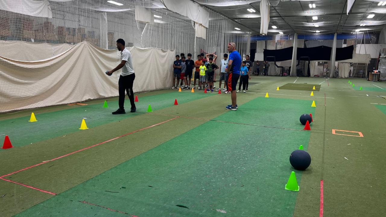A group of young cricket players attentively watch their coaches during a structured indoor training session. The session includes agility drills with colorful cones and weighted balls, emphasizing skill development, coordination, and confidence-building. The focused expressions of the children highlight their determination to master new cricket techniques. Through disciplined training and overcoming challenges, these young athletes are developing both self-belief and mental resilience, preparing them for success on and off the field