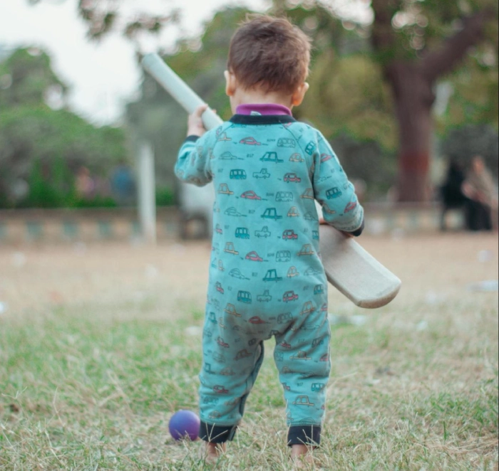 A young child in a playful, printed jumpsuit holds a cricket bat while standing on a grassy field, ready to take a swing at a small purple ball. The child's stance reflects curiosity and early engagement with the sport, highlighting the benefits of introducing cricket at a young age. With an open outdoor environment and natural surroundings, this image captures the essence of early childhood development through cricket, fostering hand-eye coordination, motor skills, and cognitive growth
