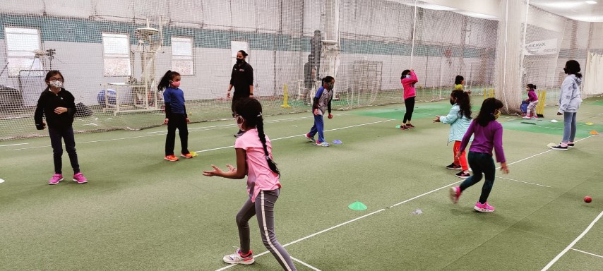 Young girls participating in a fielding practice session inside an indoor cricket training facility. They are engaged in catching and throwing drills, focusing on improving hand-eye coordination, reflexes, and accuracy. Cones are placed around the field to guide movement and positioning. A coach observes in the background, ensuring proper technique and engagement in the session.