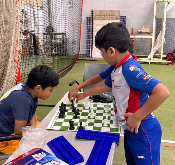 Two young boys engage in a chess game at a table inside an indoor cricket training facility. One boy, dressed in a CricMax training uniform, makes a strategic move while the other watches intently. The table also has a Rummikub game set, reinforcing the theme of blending structured learning with play. This setting highlights the balance between disciplined training and playful engagement, ensuring that children remain excited about learning and developing skills beyond just cricket.
