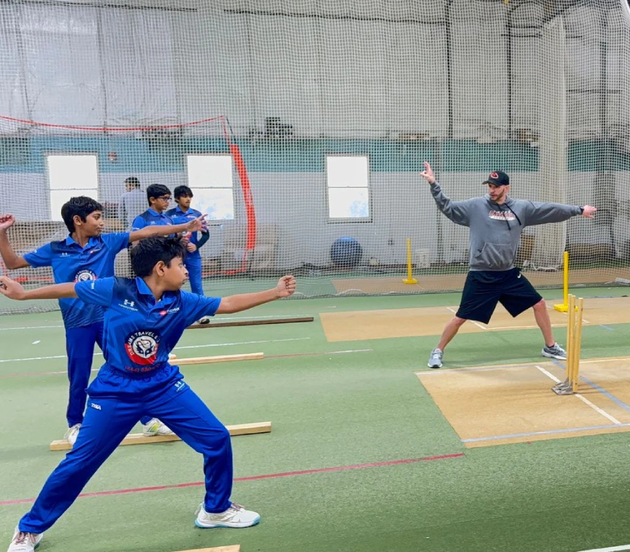 A professional cricket coach demonstrating a bowling technique to young players in an indoor net facility. The players mimic the movement to refine their form and accuracy.