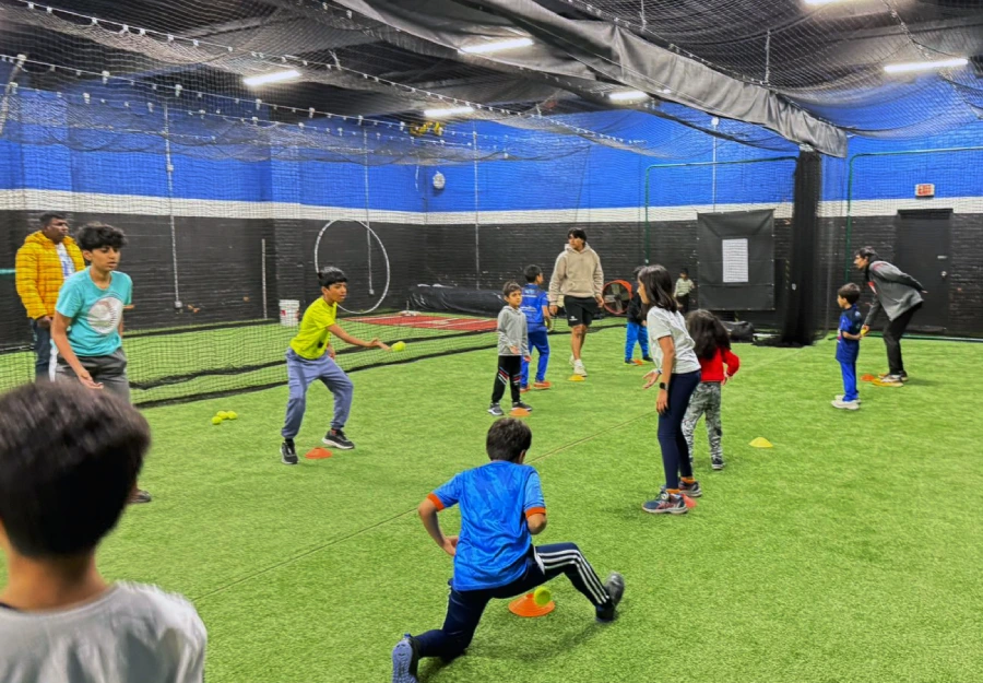 A group of children participating in a structured cricket drill session inside an advanced indoor training facility. Coaches are guiding them through fundamental exercises for fielding and reaction time.