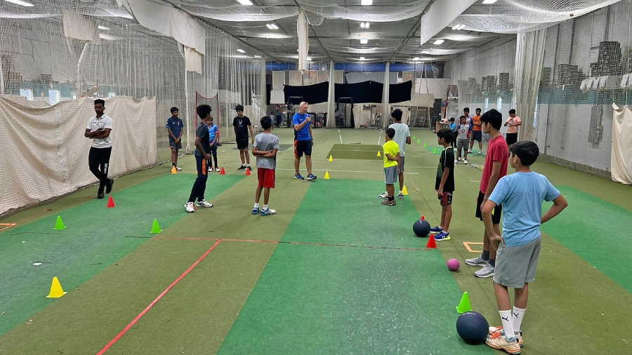 CricMax players in team uniforms discussing strategies and receiving instructions from their coach during a practice session in an indoor cricket training facility.