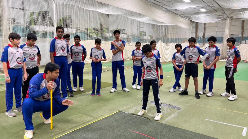 A group of young cricket players attentively listening to a coach during a training session at CricMax’s indoor facility. The coach is demonstrating a technique while the players observe in matching team uniforms.