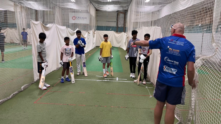 A group of young cricket players attentively listen to their coach during a personalized training session at CricMax's indoor facility. The coach, wearing a sponsor-branded shirt, gestures towards the group, providing hands-on guidance to refine their skills. The players, dressed in a mix of training gear and cricket uniforms, hold their bats as they prepare to engage in structured drills. This image highlights CricMax's commitment to individualized coaching, blending technical training with engaging sessions to help young athletes develop confidence and mastery in cricket