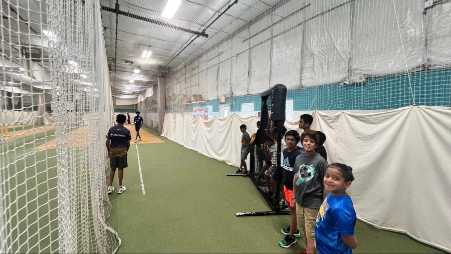 Young cricketers lined up inside an indoor cricket facility, watching a peer practice bowling in a net lane, with training nets, wickets, and coaching equipment visible in the background.