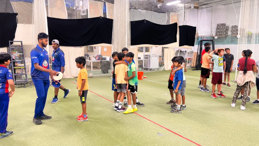 Group of young cricketers participating in a coaching session at an indoor cricket facility, guided by instructors in blue uniforms, with kids engaging in team-based drills and activities.