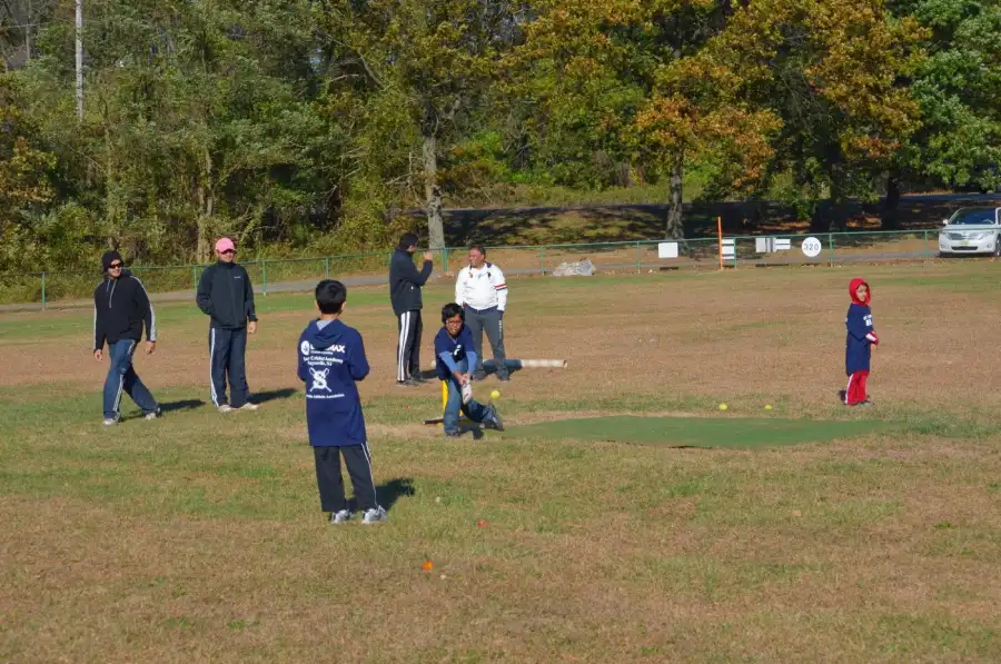 Young children participating in a cricket coaching session on an outdoor field, surrounded by attentive coaches and parents. One child takes a swing at the ball while others watch and engage, capturing the fun, supportive, and skill-building environment offered by CricMax.