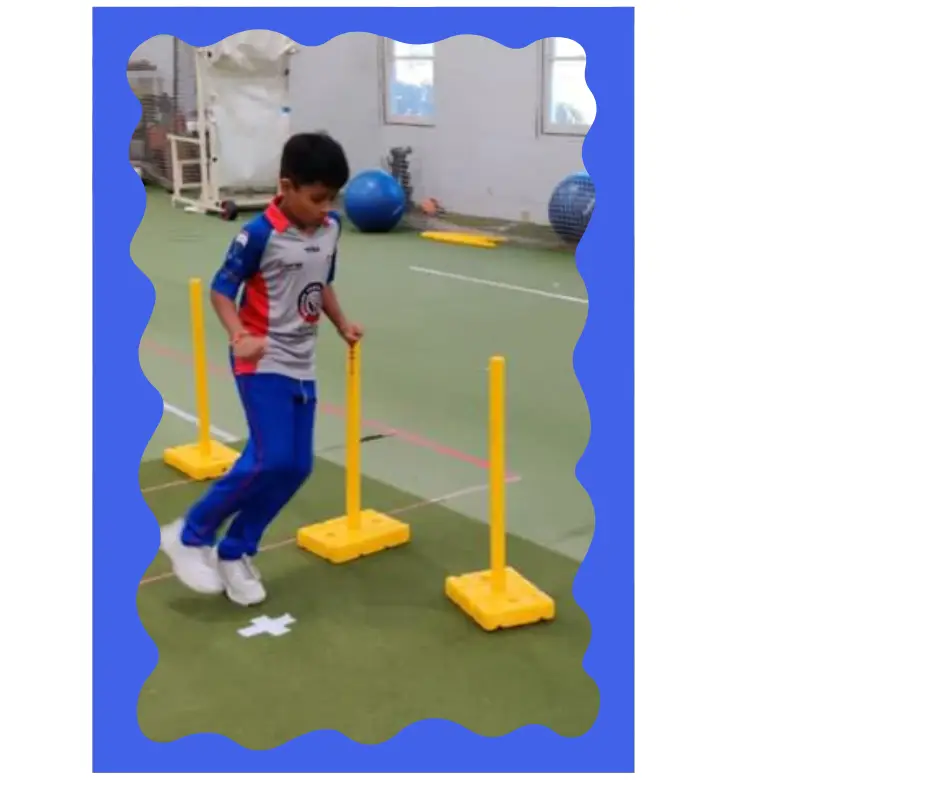 A young boy in cricket gear practices footwork and agility drills by jumping over mini hurdles during an indoor training session, focusing on decision-making and field movement awareness.