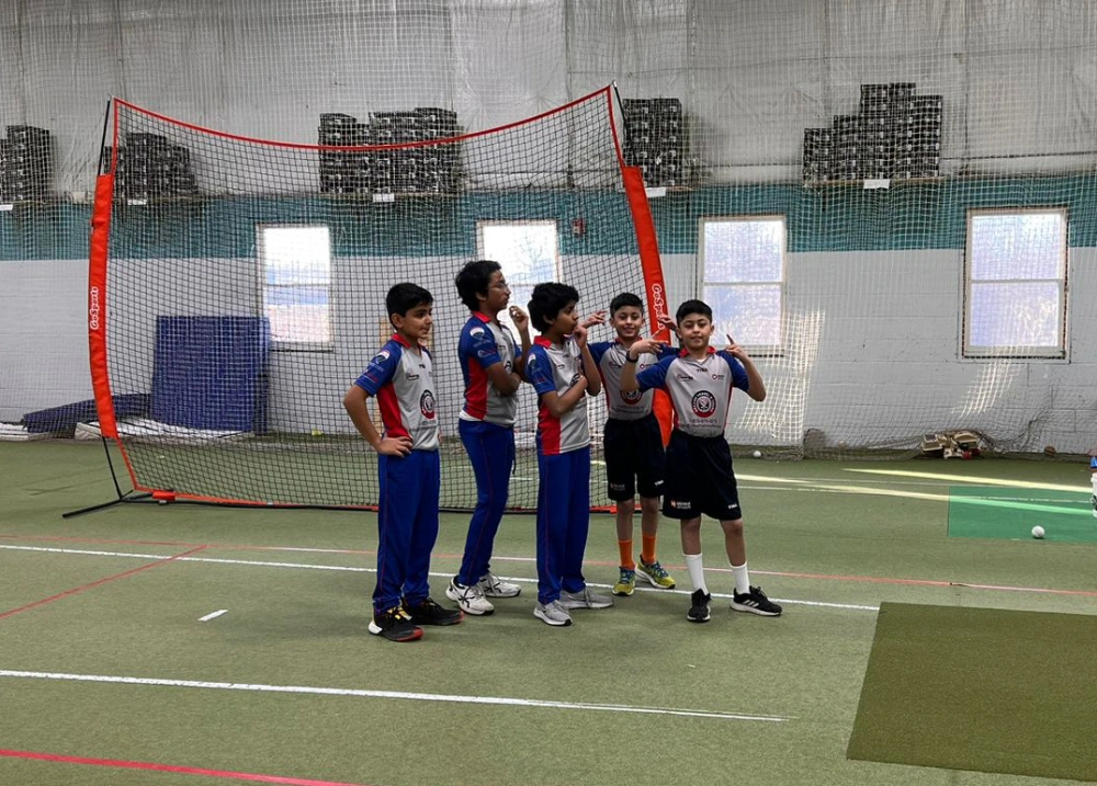 A group of young cricket players smiling and posing together in an indoor training facility. They wear matching uniforms and stand confidently near a coach, reflecting team spirit, friendship, and growing confidence through cricket.
