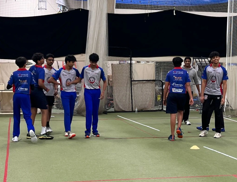 Teen cricket players in an indoor training facility listening to their coach and preparing for practice. The group shows focus, discipline, and respect during the session, highlighting the character-building aspects of cricket through structured teamwork and coaching.