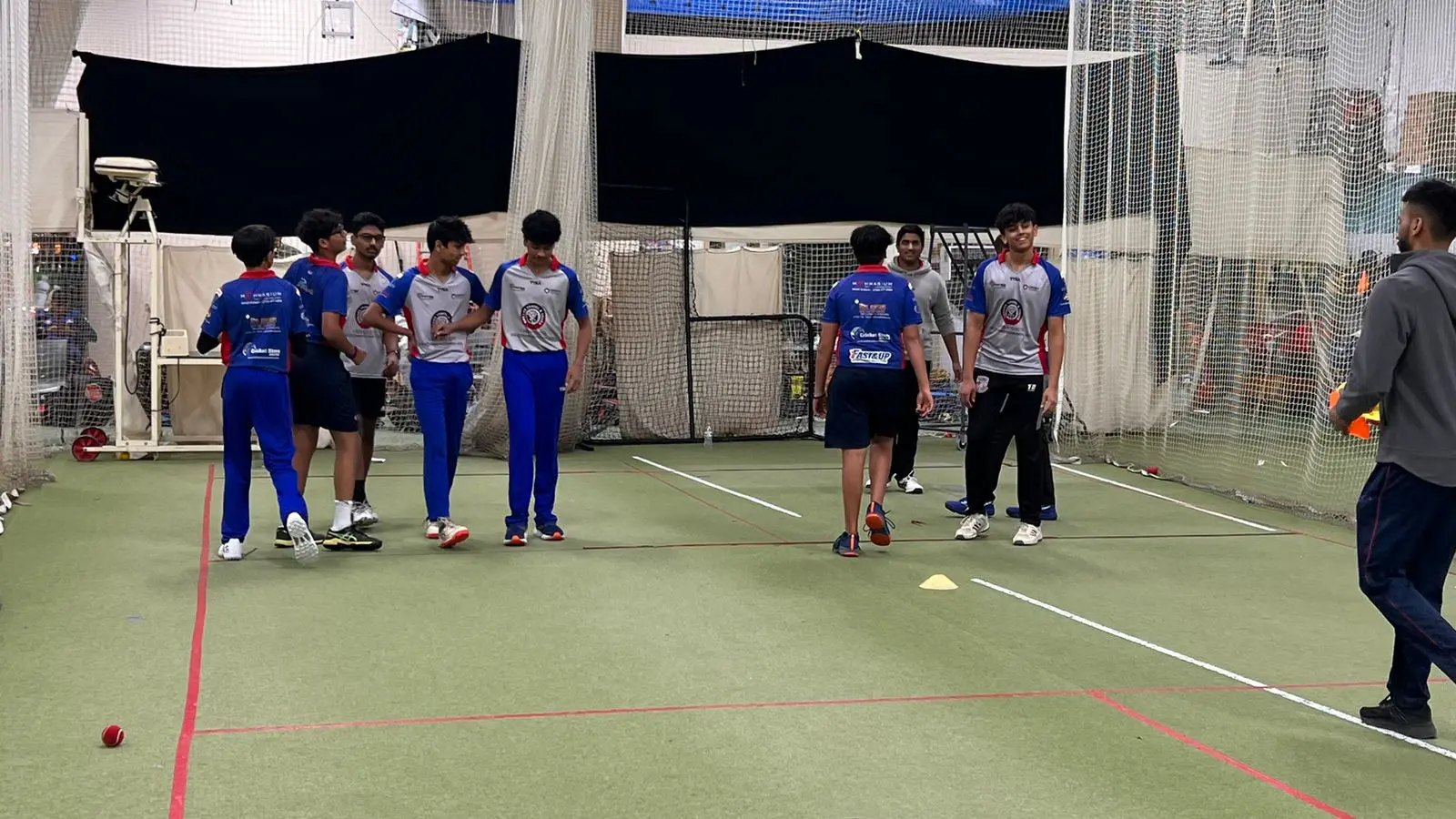 Young cricket players wearing proper safety gear, including team uniforms and athletic footwear, participate in a supervised session at the CricMax indoor facility. Coaches observe closely as players interact and prepare for drills, with netting and marked turf ensuring a safe, controlled environment—reflecting the program’s strong emphasis on injury prevention and certified instruction.
